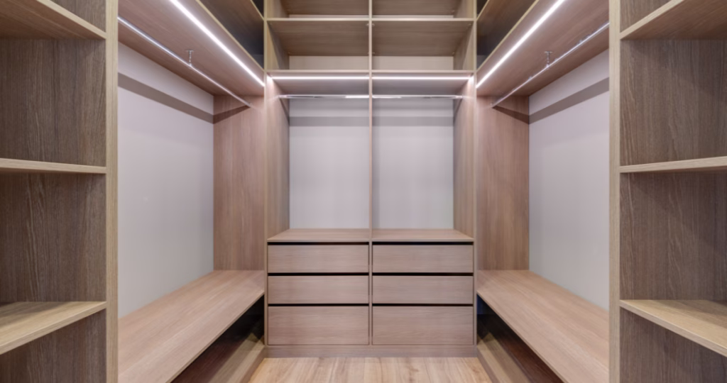 A newly installed, unfurnished floor-to-ceiling bespoke library wall in light timber. The architectural joinery features custom open shelving, integrated cabinetry drawers, and a backlit glass display niche, showcasing the raw structure and geometric precision of custom millwork.
