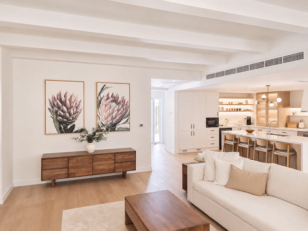 Bright open living area featuring a rich dark wood side credenza, framed botanical protea prints, light wood floors, and a view leading into the open kitchen.