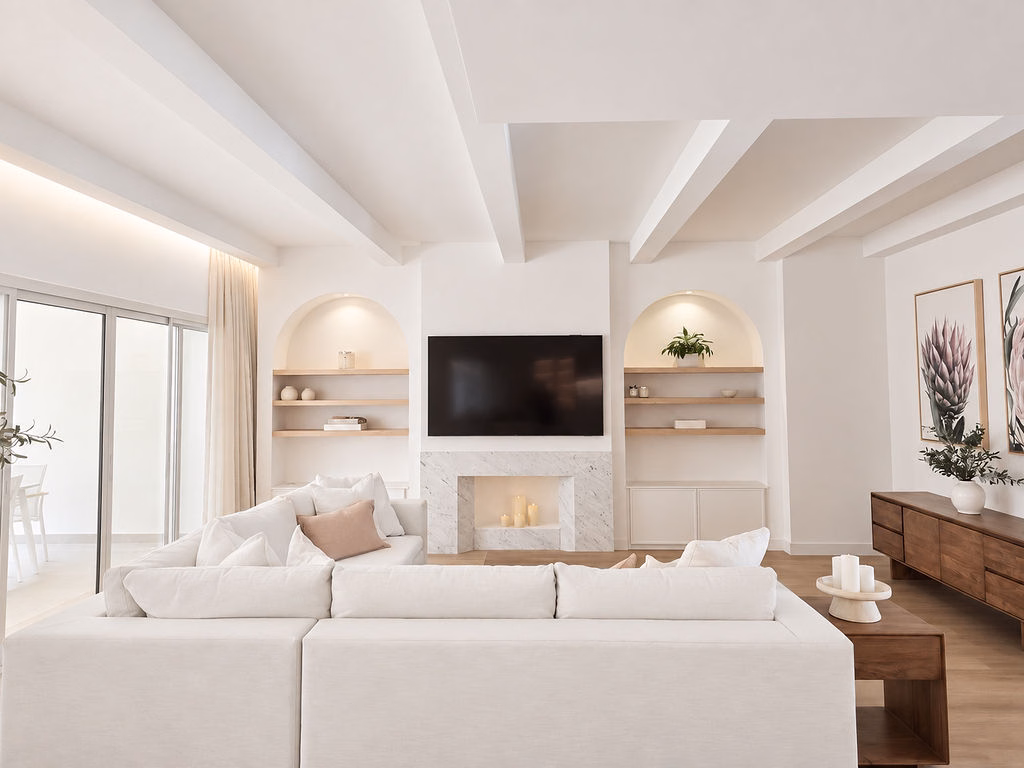 Centred view of an open-plan living room with a cream sectional sofa, marble fireplace, built-in recessed arched shelving, and white exposed ceiling beams.