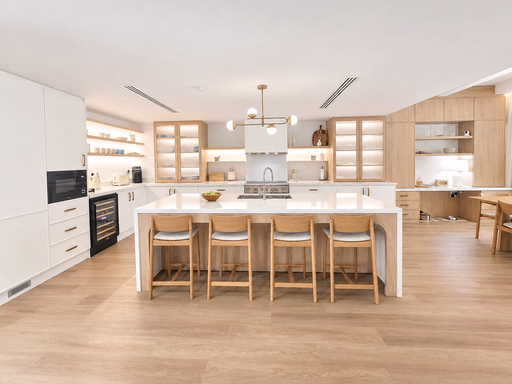 Front perspective of a large white kitchen island with four wooden counter stools, brass pendant light fixture, and floating shelves.