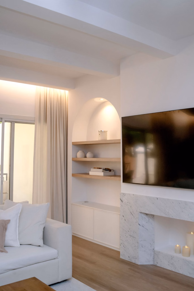 Vertical detailed view of an illuminated arched wall niche with custom wooden shelves, marble fireplace mantle, wall-mounted TV, and white beam ceiling.