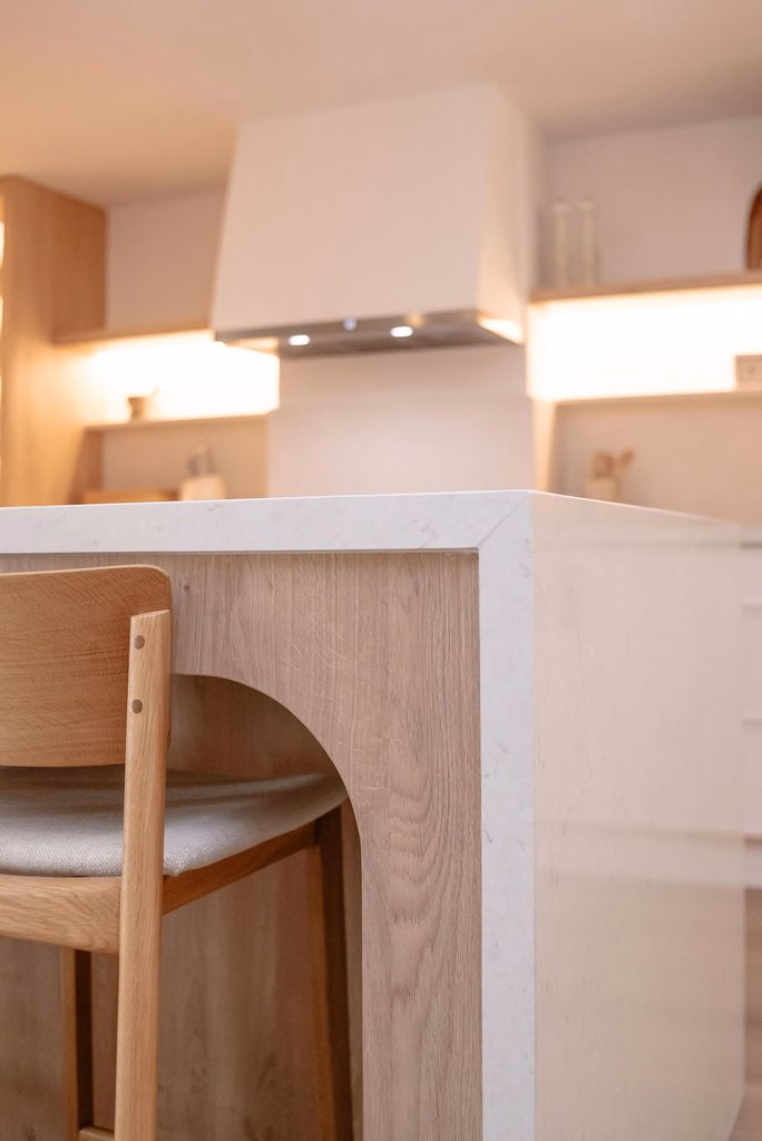 Close-up detail of a white quartz waterfall countertop island with custom light oak recessed arched leg framing and a wooden bar stool.