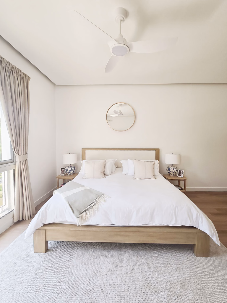 Master bedroom designed in neutral tones with a warm light-wood bed frame, textured area rug, round wall mirror, and porcelain table lamps.