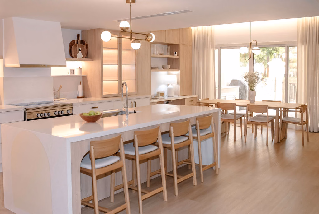 Modern open-plan kitchen featuring a custom ribbed kitchen island with wooden bar stools, soft globe lighting, and an adjoining wooden dining table with sheer curtains.
